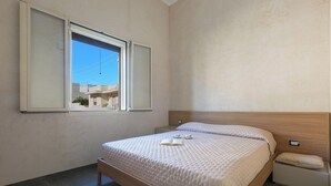 Een slaapkamer met een bed, nachtkastje, raam en uitzicht op gebouwen.