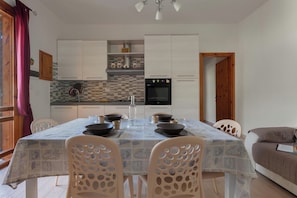 A dining area with a table set for four, surrounded by white chairs with circular cutouts. A kitchen with white cabinets and a tiled backsplash is visible in the background. - Villino Pascoli by BarbarHouse (Torre dell'Orso)