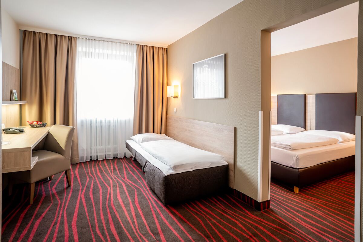 Una habitación de hotel con cama, escritorio, silla, lámpara y ventana con cortinas.