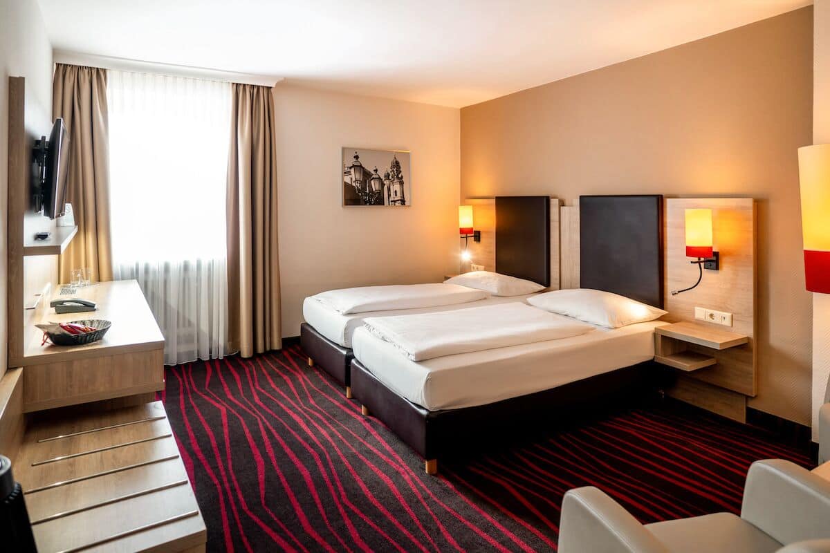 Ein Hotelzimmer mit zwei Betten, einem Schreibtisch mit Stuhl, einem Fernseher und zwei Wandlampen.