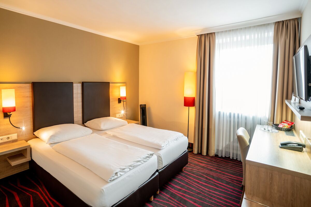 A hotel room with a double bed, bedside lamps, a desk with a TV, and a large window with curtains.