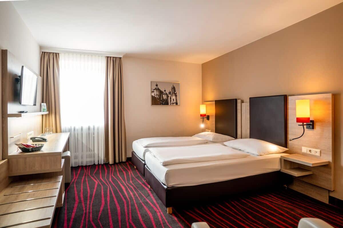 Ein Hotelzimmer mit zwei Betten, einem Schreibtisch, einem Stuhl, einem Fernseher und einem Fenster mit Vorhängen.