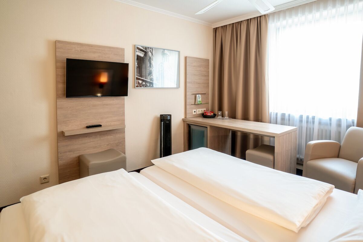 Ein Hotelzimmer mit zwei Betten, einem an der Wand befestigten Fernseher, einem Schreibtisch mit Stuhl und einem Fenster mit Vorhängen.