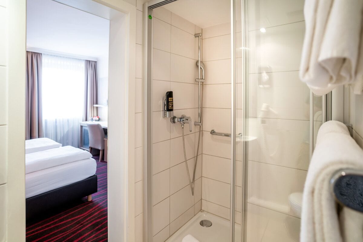 Quarto de hotel com chuveiro envidraçado, cama com carpete listrado em vermelho e preto, e banheiro branco com toalha pendurada na parede.