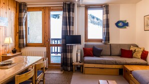 A cozy room with a wooden dining table, a sofa with cushions, a television, and a view of a snowy landscape through the windows. - 2-room apartment near slopes - 5 people (La Plagne Montchavin Les Coches)