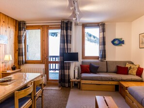 A cozy room with a wooden dining table, a sofa with cushions, a television, and a view of a snowy landscape through the windows. - 2-room apartment near slopes - 5 people (La Plagne Montchavin Les Coches)