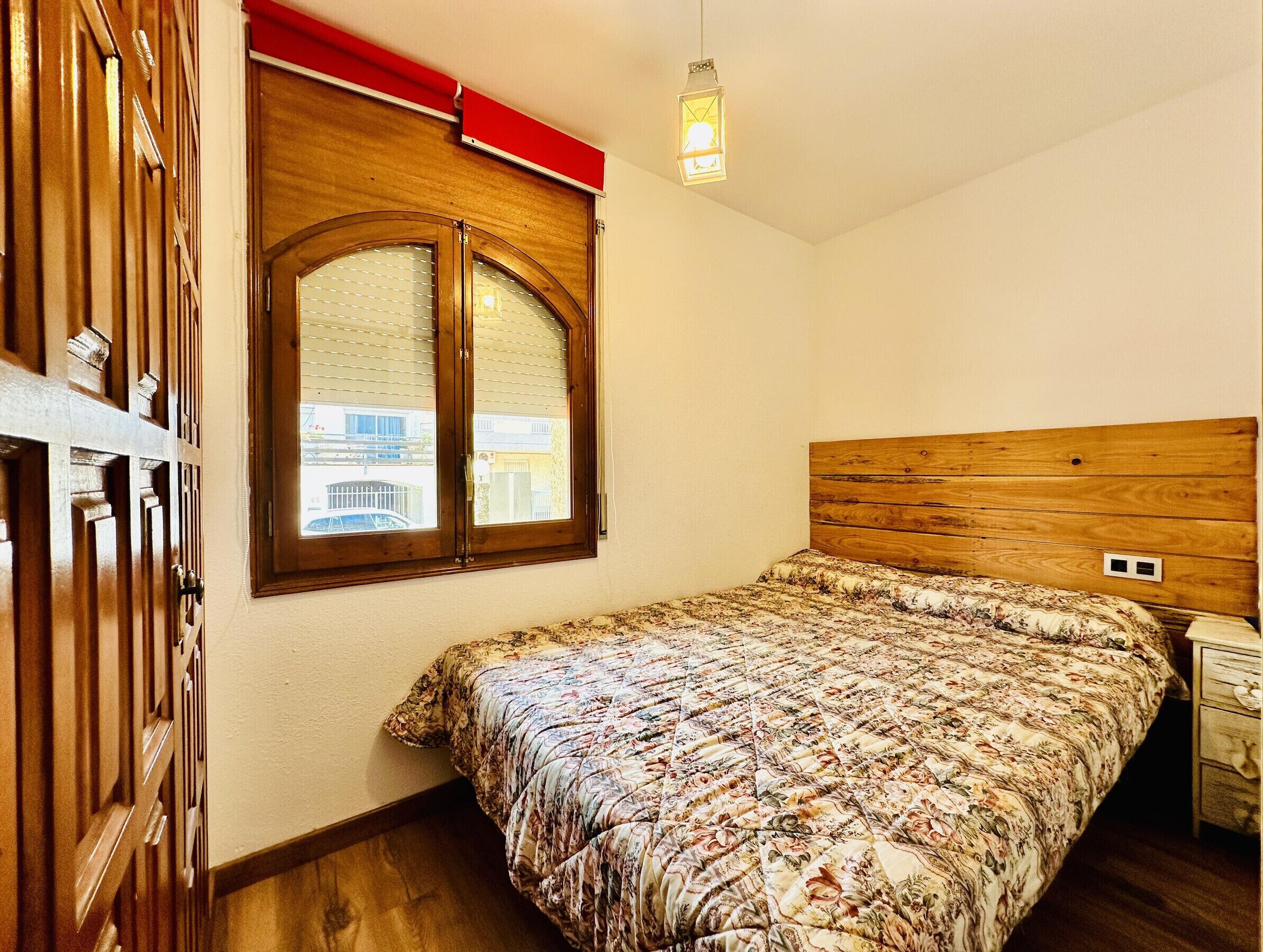 A bedroom with a wooden headboard, a floral bedspread, a window with blinds, and a hanging light fixture.