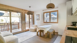 A modern kitchen with a dining area, large windows, and a sliding glass door leading to an outdoor deck. - Charming T2 with cabin 50m from slopes for 4-5 pers (Les Gets)