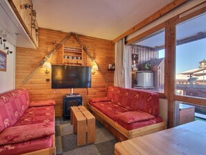 A cozy cabin interior with wooden walls, a large TV, two red sofas, a wooden table, and a window overlooking a snowy landscape. - Central studio with balcony in Belle Plagne, 4 pers (Belle Plagne)