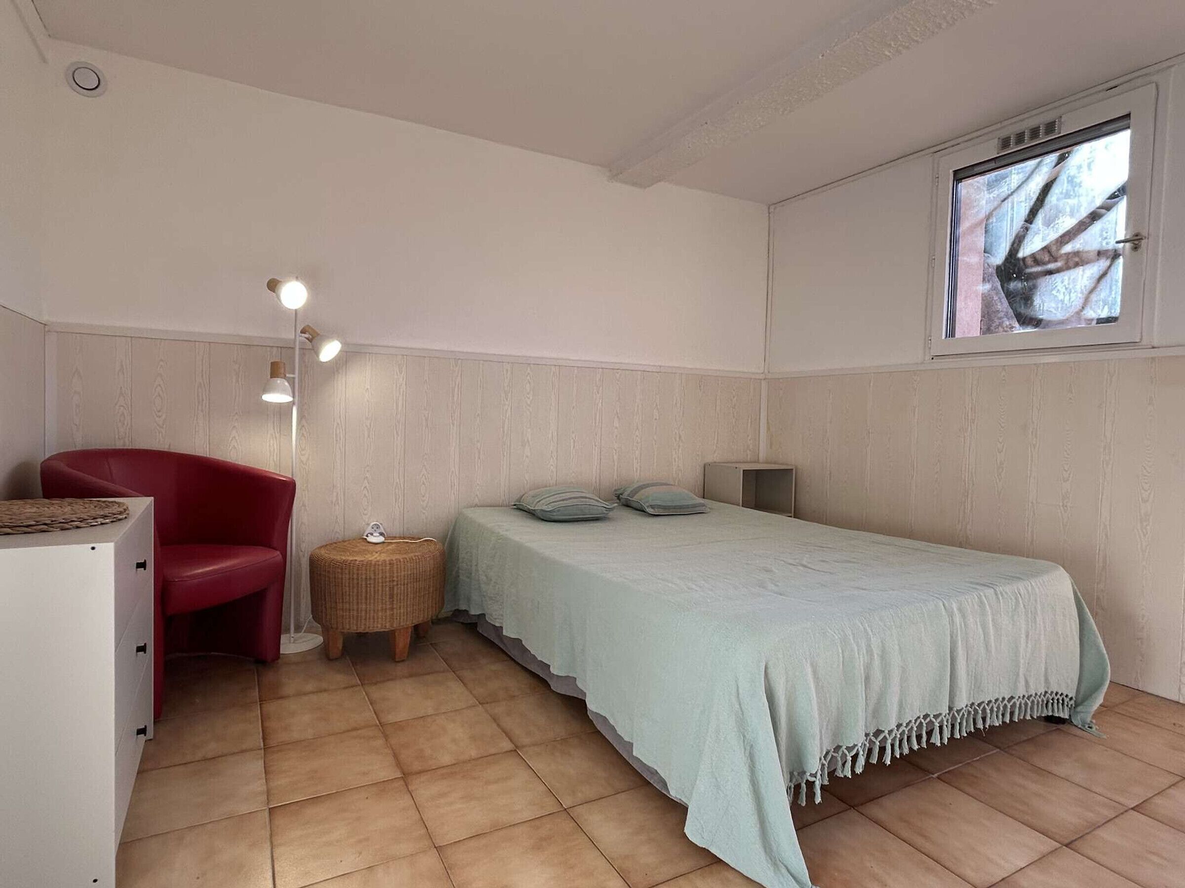 A bedroom with a bed, a red chair, a small round table, a lamp, and a window with a view of trees.