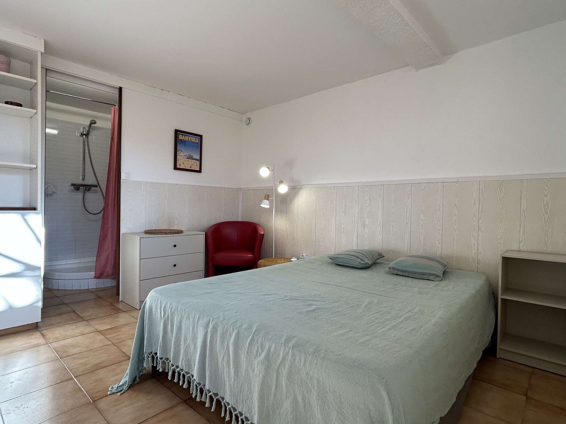 A bedroom with a bed, a red chair, a white dresser, a wooden wardrobe, a shower, and a wall-mounted lamp.