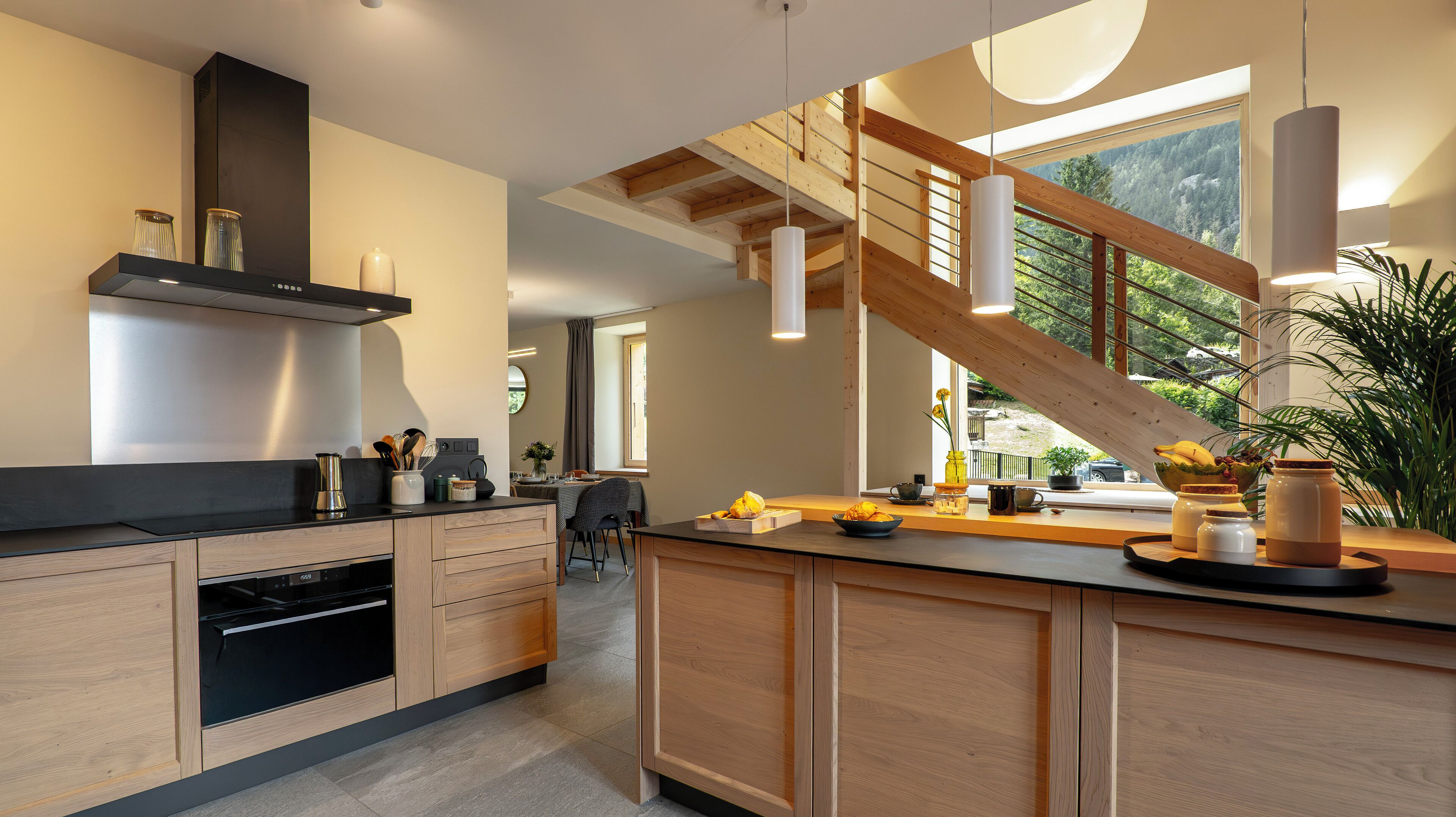 A modern kitchen with wooden cabinets, a built-in oven, and a large island with a sink. There is a staircase with a wooden railing leading to an upper level.