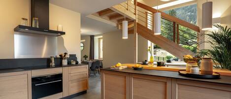 A modern kitchen with wooden cabinets, a built-in oven, and a large island with a sink. There is a staircase with a wooden railing leading to an upper level.