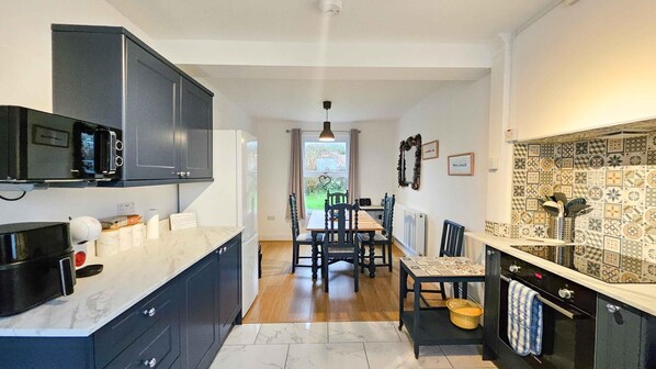 A modern kitchen with dark cabinetry, a marble countertop, and a dining area with a wooden table and chairs. - Brynview - 3 Bedroom Holiday Home - Reynoldston (Swansea)