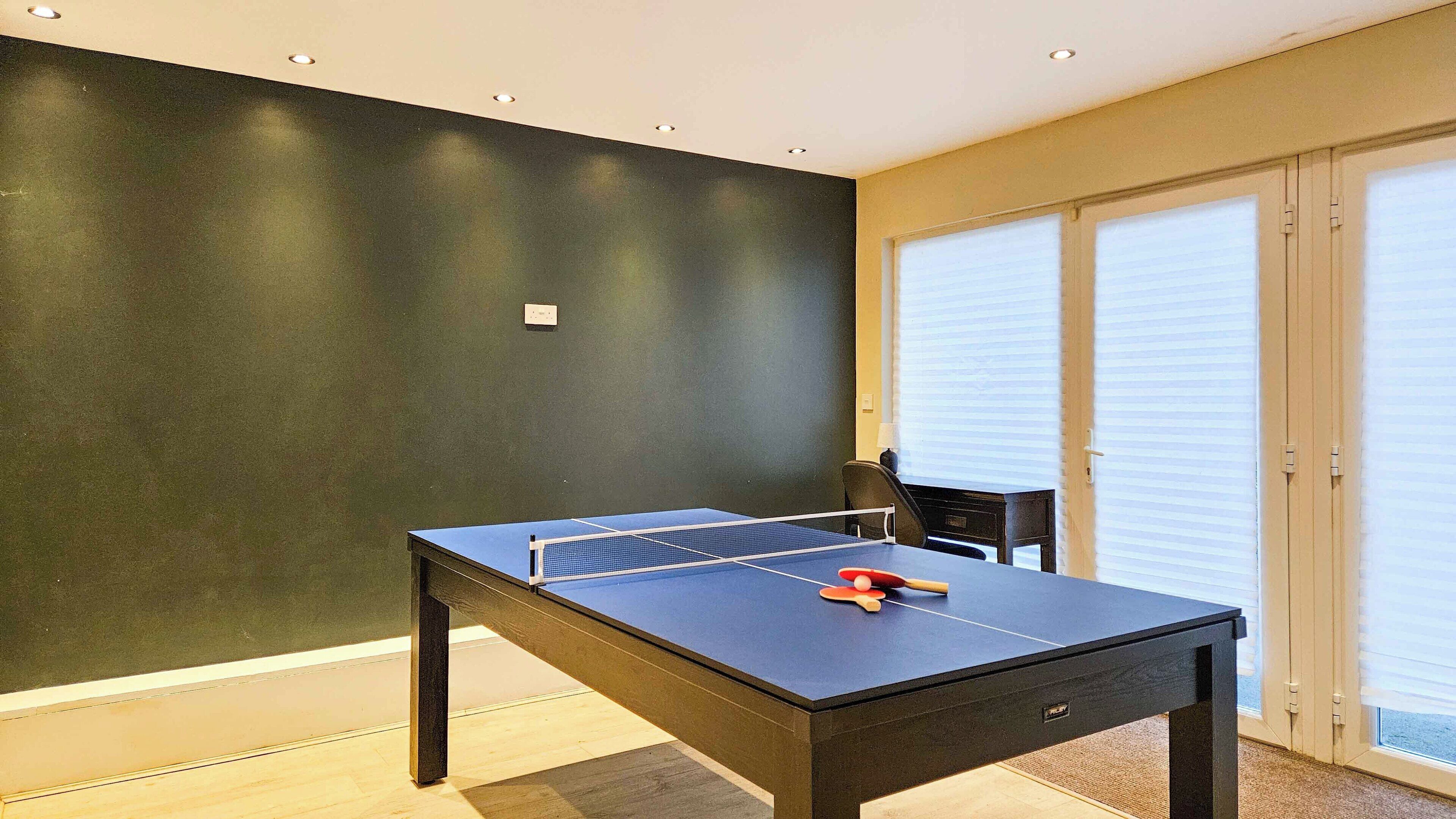 A ping pong table in a room with a dark wall, a door with blinds, and a chair.