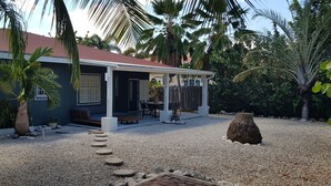 Een huis met een overdekt terras, een grindpad en omliggende tropische begroeiing.