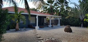 Ein Haus mit überdachter Terrasse, einem Schotterweg und üppiger tropischer Bepflanzung.