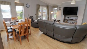 A modern living room with a dining area, grey leather sofa, wooden dining table, and a flat-screen TV mounted on the wall. - Holly Lodge (Holywell)