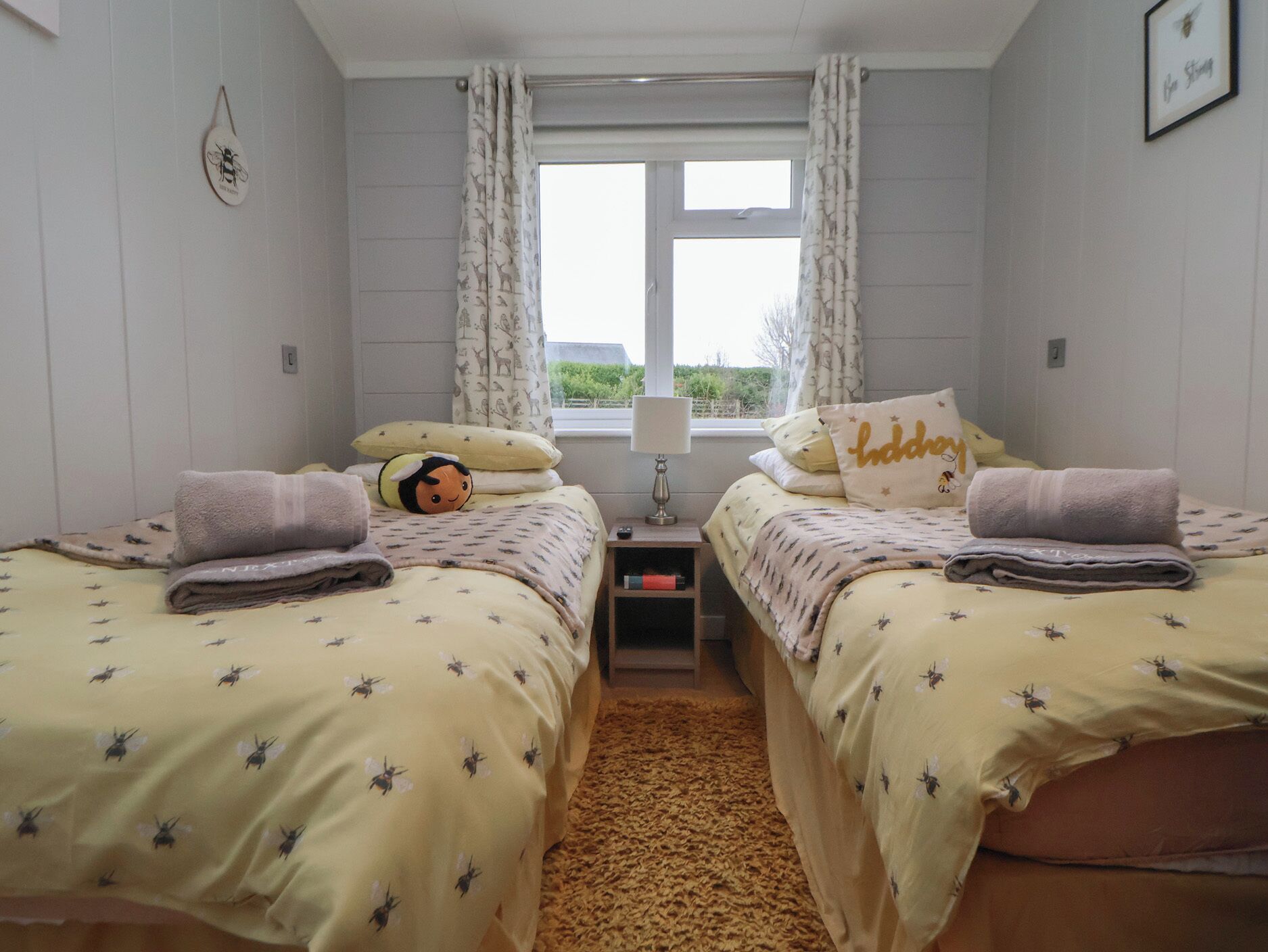 Ein Zimmer mit zwei Betten, jeweils mit gelber Bettdecke mit Bienenmuster, einem kleinen Nachttisch mit Lampe und einem Fenster mit Vorhängen.