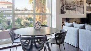 A modern living room with a round table and chairs, a sofa, a large window with a view of buildings and trees, and a framed picture on the wall. - New 2 Bedrooms, 2 Bath Apt in Central Glyfada, Fast WiFi, 10Min Walk to Beach (Glyfada)