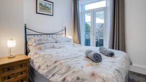A neatly made bed with floral bedding, a bedside lamp, and a wooden nightstand with drawers. - Caldey Island View - 2 Bedroom Apartment - Tenby (Tenby)