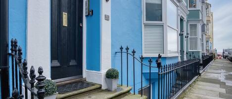 Una fila de casas escalonadas con fachadas azules y blancas, un portón de hierro negro y una pequeña planta en maceta en los escalones.
