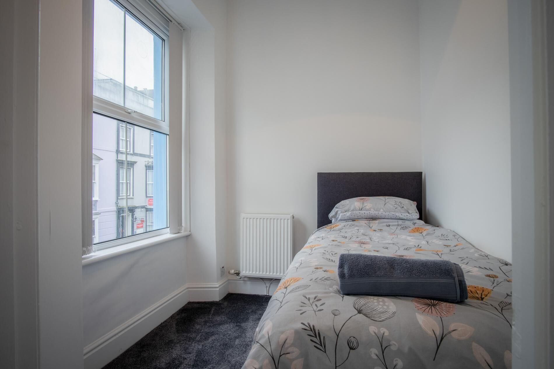 Une petite chambre à coucher, bien rangée, avec un lit simple, un radiateur et une fenêtre donnant sur les bâtiments environnants.