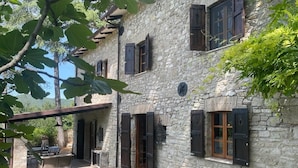 Ein Steinhaus mit zahlreichen Fenstern und Fensterläden, einer überdachten Terrasse und einer Uhr an der Wand.