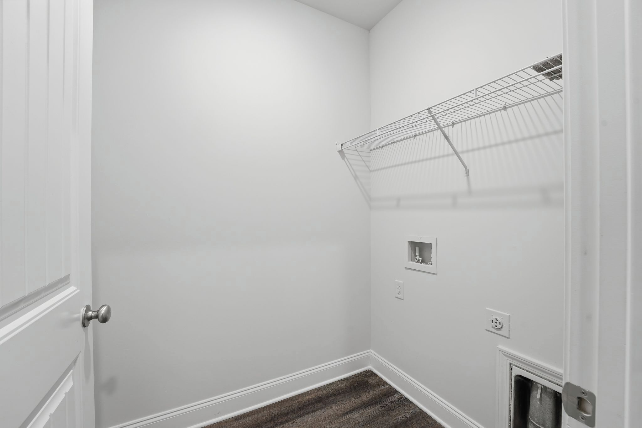 A corner room with white walls, a wooden floor, a door, a metal shelf, and a small electrical outlet.