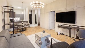 A modern living room with a large flat-screen TV, grey sofas, a coffee table, and a dining area with chairs and a small kitchen in the background. - Apartment Husarski comfort 2-bedroom apartment - swimming pool and saunas (Poznań)