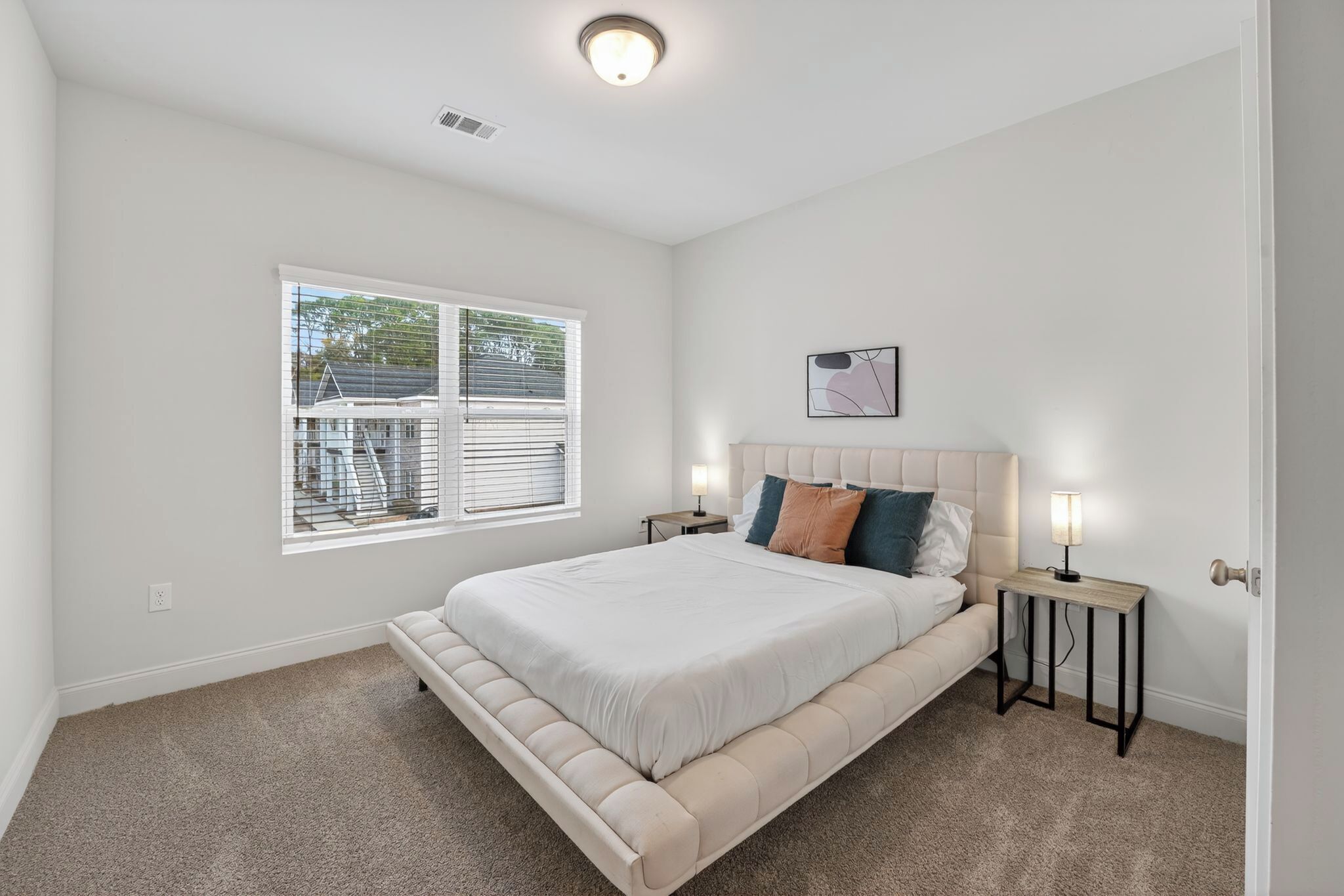 A bedroom with a large bed, two bedside tables with lamps, a window with blinds, and a ceiling light.