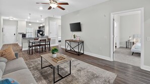 Una sala de estar moderna con ventilador de techo, un comedor con mesa y sillas, una cocina al fondo y un televisor montado en la pared.