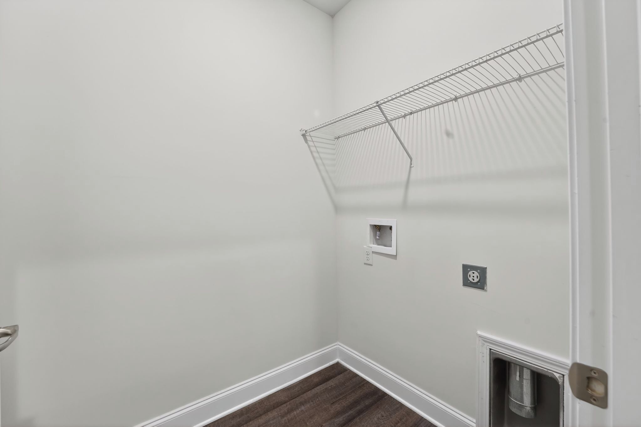 A corner of a room with a white wall, a metal wire shelf, a small electrical outlet, and a door with a metal handle.