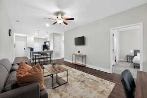 Un salon moderne avec un canapé, une table basse et un ventilateur de plafond. On aperçoit, à l’arrière-plan, un coin cuisine avec une table à manger et des chaises.
