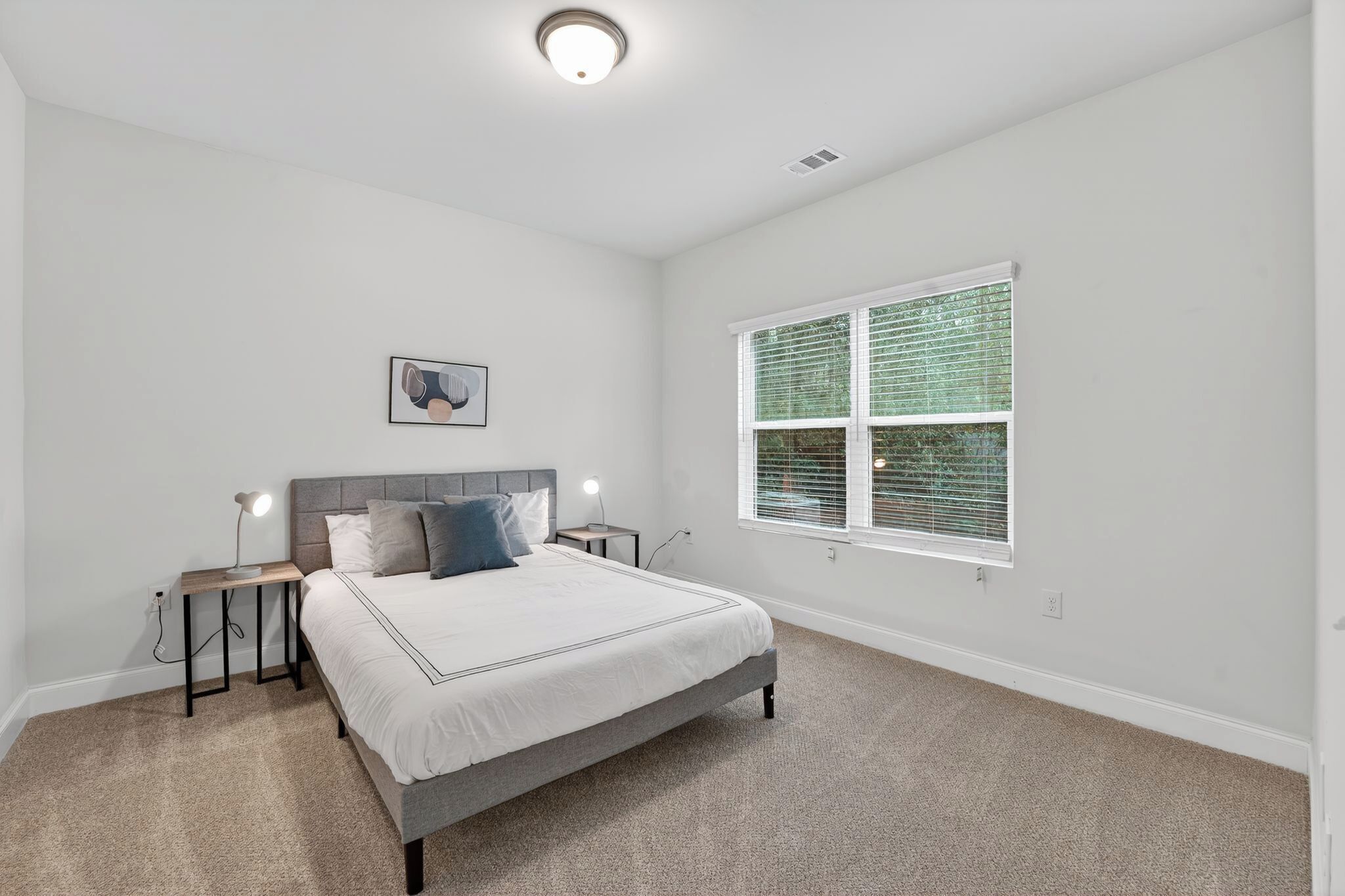 A bedroom with a bed, nightstands, a wall-mounted light, a window with blinds, and a ceiling light.