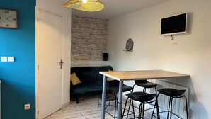 Een klein leefruimte met kitchenette, een blauwe accentmuur, een muur met steenpatroon, een houten eettafel met barkrukken, en een aan de muur bevestigde televisie.