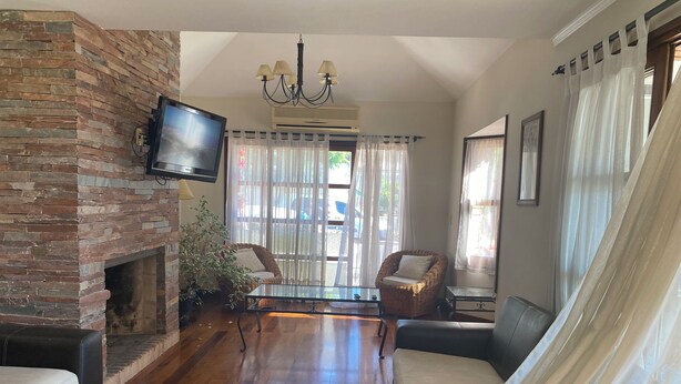 A living room with a brick fireplace, a flat-screen TV mounted above it, a chandelier, wicker furniture, and a large window with sheer curtains. - HOTEL AYRES (Colonia del Sacramento)