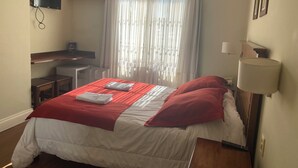 A hotel room with a bed, a desk, a chair, a TV, an air conditioner, and a window with curtains. - HOTEL AYRES (Colonia del Sacramento)