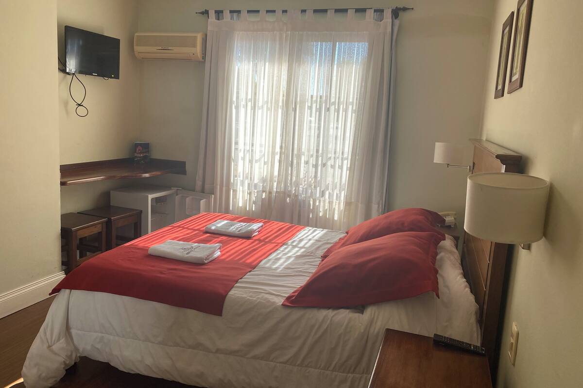 Habitación de hotel con cama, escritorio, silla, televisión, aire acondicionado y ventana con cortinas.