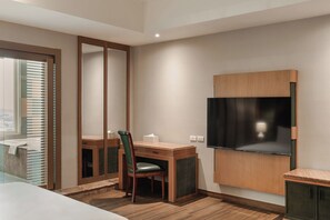 A modern hotel room with a wooden desk, a chair, a large mirror, a flat-screen TV mounted on the wall, and a window with blinds. - MUMU JIAO-XI (Jiaoxi)