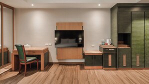 A compact living space with a wooden desk, a green chair, a flat-screen TV mounted on the wall, and a kitchenette area with cabinets and a countertop. - MUMU JIAO-XI (Jiaoxi)