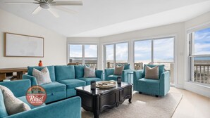 Amplia sala de estar con sofás color turquesa, una mesa de centro negra y ventanas grandes con vistas a la playa.