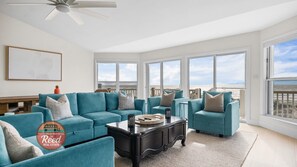 Ein geräumiges Wohnzimmer mit türkisfarbenen Sofas, einem schwarzen Couchtisch und großen Fenstern mit Blick auf den Strand.