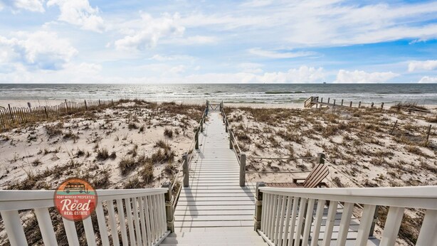 Un paseo marítimo de madera que conduce a la playa, rodeado de arena y vegetación escasa.