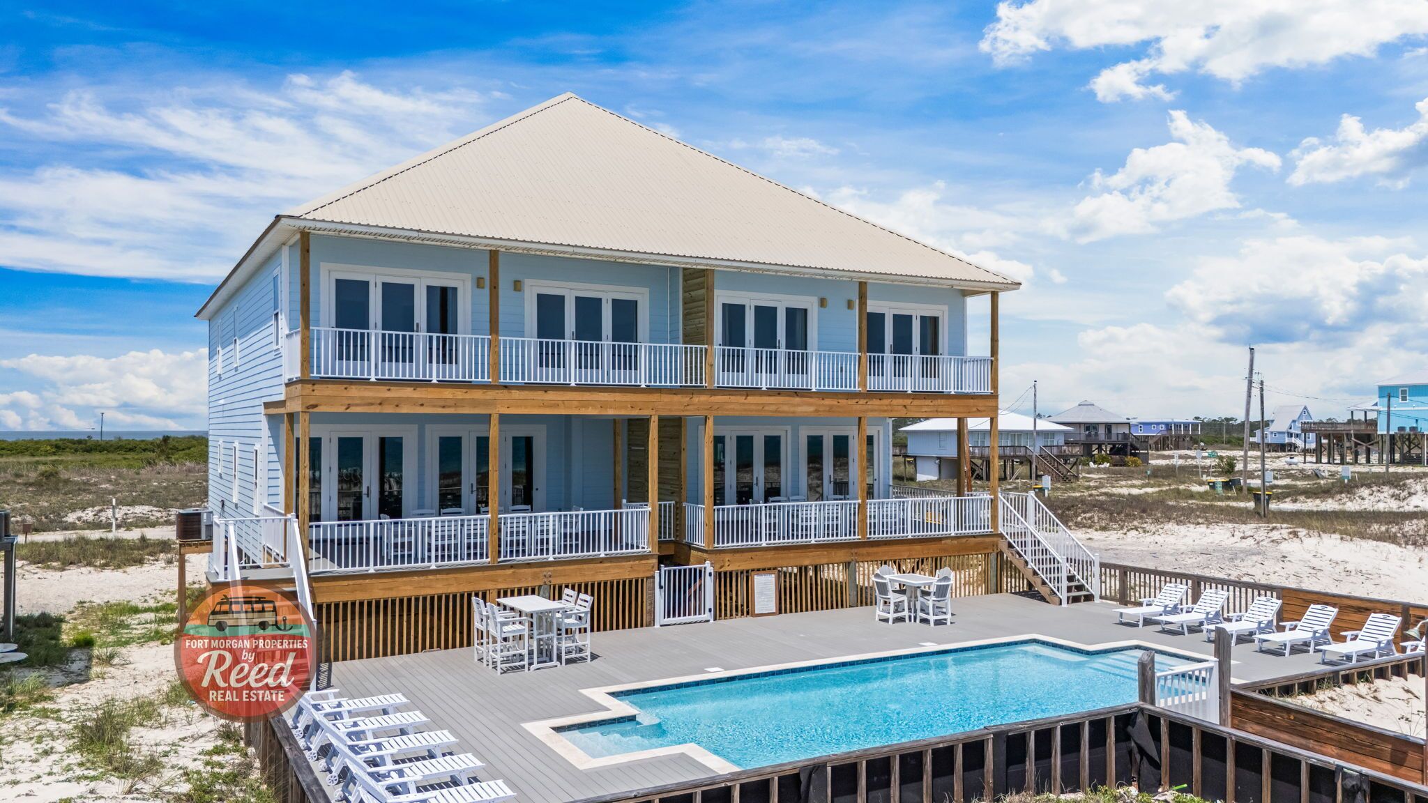 A two-story beachfront house with a pool, deck chairs, and a wooden deck.