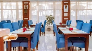 A hotel dining area with blue upholstered chairs, wooden tables, and large windows with sheer curtains. - König Hotel (Pécs)