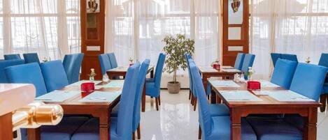 Un comedor de hotel con sillas tapizadas en azul, mesas de madera y ventanas grandes con cortinas translúcidas.