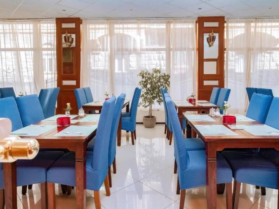 Un comedor de hotel con sillas tapizadas en azul, mesas de madera y ventanas grandes con cortinas translúcidas.