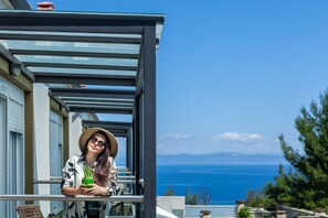 Een balkon met een helder uitzicht op zee en bergen, een persoon met een drankje, en een moderne architectuurstijl.
