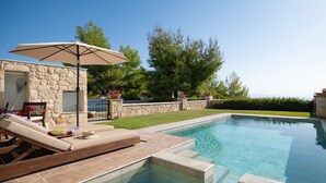 Una piscina con muretto in pietra, una zona relax con una sedia e un ombrellone, e vista su alberi e cielo sereno.